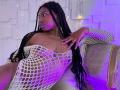 QueenPerlaa with dark skin, long black braids, wearing a white fishnet dress, exposing her tits and pussy, sitting on a beige couch, purple and white lights in the background.