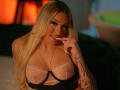 CalistaRenzi with long blonde hair, smiling, finger near her lips, wearing a black and sheer top, showing her big tits, tattooed arm, and gold necklace. Warm, orange-lit background.