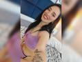 MartinaRey with long black hair, purple bra, and black thong, smiling, showing a butterfly tattoo on her right arm, in a bed with geometric-patterned sheets.