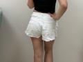 Tilliana's white, frayed butt shorts, showing her ass, with hands on hips, wearing a black top, standing against a plain wall, photographed from behind.