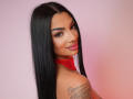 NatalieDelices has long black hair, red lipstick, and a red scarf. She's looking over her shoulder with a smile, showing a tattoo on her right shoulder.