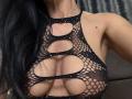 AndraD with black fishnet top, showing large boobs through cutouts, black hair, and a small mole on her right shoulder. She's wearing a black, sparkly, halter-style top.