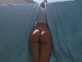 JuneLaFrenchie's ass is exposed, standing with blue curtains open, wearing a white top, her tan skin and butt highlighted in the sun, from behind, in a photo.
