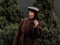 VirgieCrotts in a brown coat, black hat, holding a phone to her ear, with green shrubs in the background, looking slightly annoyed. She has long brown hair and nails.