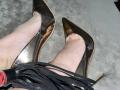 GeraNia's pale legs in shiny black stiletto heels, with a black leather flogger's straps in the bottom left corner. Her feet are pointed, showing the heel's reflection.