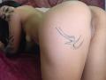 LuciadelaVega, naked, on all fours with her ass up, showing a bird tattoo on her right buttcheek. Her black hair, tattoos on her arm, and visible pussy are highlighted.