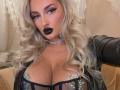 MademoiselleBella with platinum blonde curls, black lipstick, choker, and black leather jacket, showing big tits in a sheer black top with glittery pasties.