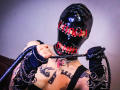 AmySpider wearing a shiny black latex mask with red eye holes, holding a black leather whip, has tattoos on her chest, and is in a black top.