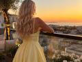 EliseMonet with long, wavy blonde hair in a yellow, backless dress, standing on a balcony, leaning on a glass railing, looking at a sunset over a city.