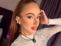 MarissaFex with brown ponytail, wearing white lace top, diamond earrings, and necklace, touching head, full lips, brown eyes, subtle makeup, looking at camera, purple curtain background.
