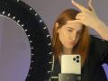 Malvina Hauenstein, con cabello rojo, haciendo un gesto de paz, sosteniendo un teléfono inteligente, frente a un aro de luz negra con luces LED blancas, vistiendo una camisa negra..