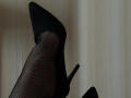 Keiri's leg in black fishnet stockings and pointed black high heels, with a stiletto heel, showing a sexy, elegant, and confident vibe.