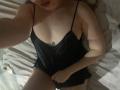 JannaCorsaro in a black, sheer, spaghetti-strap top, exposing small, round tits with a tiny, blue, flower tattoo above. She's lying on a bed, covering her pussy.