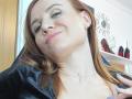 HeartlessQueenn with light skin and red hair, smiling with a slight tilt, wearing a black shirt, pink lips, subtle makeup, and a thin necklace, with a gentle hand on her chest.