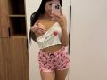 AuraXo with long black hair, white crop top revealing one boob, pink strawberry shorts, holding phone, smiling, showing tattoo on right hand, standing in front of mirror.