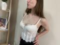StellaNoir with long brown hair, pouty lips, wearing a white lace bustier that shows her small tits, hands on hips, black pants, silver necklace, in a softly lit room.