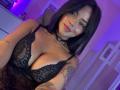 MajoJones with large boobs, lace black lingerie, choker, tattoos, and long dark hair, smiling with pink lipstick, purple lighting.