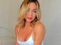 EllisaWoman with blonde hair, red lipstick, white tank top, showing her tits, fair skin, smiling, sitting on a bed with white sheets.