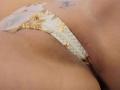 Close-up of ElixirShanna's crotch showing a white lace thong with gold floral embroidery. The thong is slightly wedged between ElixirShanna's slightly parted, smooth, tan-colored labia. A small, dark tattoo is visible above the thong on ElixirShanna's right hip. The lace has a zigzag pattern on the sides. The texture of the lace contrasts with ElixirShanna's smooth skin. The image focuses on the intimate area, with the thong and tattoo being the primary elements.