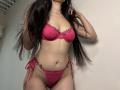 Tisia in pink lace lingerie with floral trim, showing her tits and pussy in a photo. Long dark hair, hand in hair, standing against a plain wall.