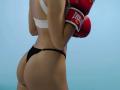 MilagrosEdmisten stands with back to camera, wearing a white sports bra and black thong, holding red boxing gloves. She has a toned, tanned body, defined buttocks. Bright blue background.