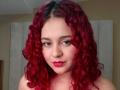 RosalieVittoria with red curly hair, pinkish skin, brown eyes, and red lipstick, looking directly at the camera with a slight smile. Her tits are not visible.