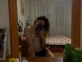 Topless GwynBartell with small tits and black panties, sitting in front of a mirror, holding a phone, taking a selfie, in a dimly lit room, with a bed behind.