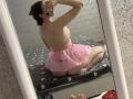 KaiaLein in pink lingerie, sitting on a bed, facing mirror, showing her ass and back, wearing a sheer pink skirt, holding a phone, dark hair in a bun.