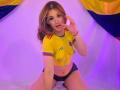ElodieGardner with glasses, yellow crop top showing small tits, red and blue trim, and black shorts, kneeling, touching glasses, pink and purple lighting, soft smile.