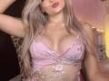 EliseMonet, avec ses longs cheveux blonds, ses lèvres roses et pulpeuses, portant un soutien-gorge en dentelle rose transparent, mettant en valeur ses seins volumineux et ronds, ornés de broderies florales..