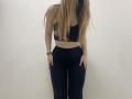 Palerianna stands against a plain white wall. She has long, straight, blonde hair covering her face. She wears a black sports bra and black high-waisted leggings. Her right hand rests on her thigh while her left hand hangs by her side. She has a black bracelet on her left wrist. The image focuses on her from the chest down, with her hair partially obscuring her face. Her skin is light. The overall composition is simple and centered.