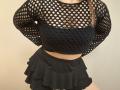 Niferttiti wearing a black fishnet crop top with visible boobs through the holes, and a ruffled black skirt, standing with hands behind her back.