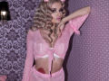MademoiselleBella with blonde curls, pink headband, sheer pink top, white lace bra, pink mesh skirt, rose tattoo on right hip, arm raised, pink nails, purple lipstick.