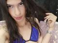 LunarisSmith with long dark hair, purple shiny bikini top, piercings in both lower lips, holding hair, silver bracelet, and pink nails, looking at camera.