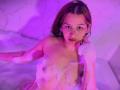Elodie Gardner, nuda, con i capelli castani bagnati, occhiali e bolle di sapone che le coprono i piccoli seni e i capezzoli rosa, seduta in una vasca da bagno illuminata di viola, che guarda verso l'alto..
