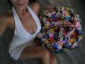 Olieri with large boobs in a white halter dress, holding a colorful bouquet, sitting on a wooden floor. Olieri's face is partially visible, with a small tattoo.