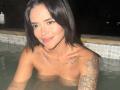 MajoJones with black hair, small tits, tattoos on chest and arm, in a pool at night, smiling, water up to her chest, looking at camera.