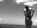 Black-and-white photo of AlexXas from behind, standing on a beach, adjusting a thong bikini, showing their ass, with a cloudy sky above and ocean in the background.