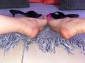 Las plantas de los pies de IrisBlair hacia arriba, los pies sobre una alfombra gris con flecos, con unos stilettos negros y un juguete sexual rosa entre ellos, sobre un suelo de baldosas. Sus dedos ligeramente separados..