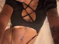 MajoJones in black strappy top with criss-cross design, showing her tits and tattoos on arms and belly, wearing thin black straps on her lower body.