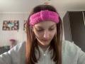 AdrianneEllinger has brown hair, wearing a pink fuzzy headband, grey hoodie, and white earbuds, looking down with a slight smile. Her tits are small and she's cute.