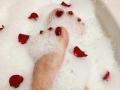 VanesaMoka's foot with shiny red-painted toenails, partially submerged in bubbly white bathwater, scattered with red rose petals.