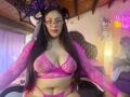 CristalZafiro in pink fishnet lingerie, showing big tits and a soft belly, with long black hair, glasses, and pink ears, sitting on a chair in a cozy room.