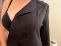 Il blazer nero di EmmaNoa, con doppie tasche, che copre il suo seno scoperto da un reggiseno nero, mostra una leggera scollatura e la sua spalla nuda. La sua pelle è chiara. Si vede un oggetto bianco..