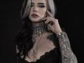 ErgoProxy with black and silver hair, wearing a lace black top, heavy chain necklaces, and a tattooed arm, touching her face with a serious expression. Boobs visible.