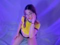 ElodieGardner, wearing a yellow tee with red stripes and black panties, sits with her hand on her cheek, blue-purple light on her, glasses on, looking down.