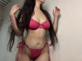 Tisia with long dark hair, wearing a pink lace bra and matching thong, hands in hair, showing her tits and pussy, standing in a room.