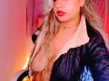 AnastaciaMonaco with long blonde hair, red lipstick, and a gold choker, wearing a black jacket with white fur, exposing her large, natural tits, in a pink-lit room.