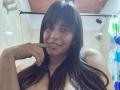 MadissonFontaine, nude, with long black hair, smiling, touching her finger to her chin, showing small moles on her cheek, in a bathroom with a shower curtain.