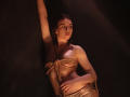 TriciaBarnes with rope bound to her tits and arms, golden light on her pale skin, dark background, arm raised, looking off to the side, slightly seductive expression.